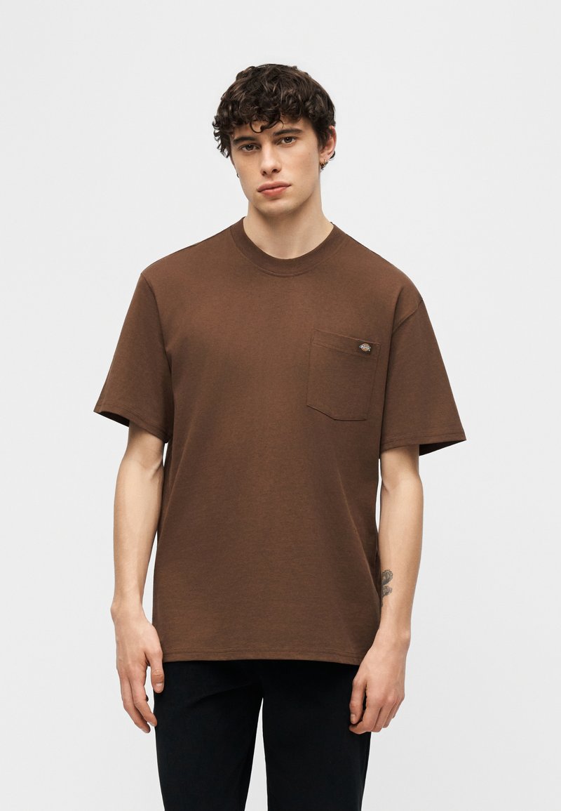 Giovane uomo con capelli ricci che indossa una maglietta marrone a maniche corte con taschino sul petto e pantaloni neri, in piedi davanti a uno sfondo bianco.