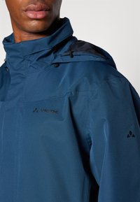 Zila ūdensizturīga jaka ar augstu apkakli, teksturētu audumu un sietu akcentiem. Iekļauj logotipu uz krūtīm un pleca, regulējamas auklas.