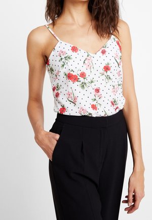 Frau in einem weißen ärmellosen Top mit rotem Blumen- und schwarzem Punktmuster, kombiniert mit schwarzen hoch taillierten Hosen, die eine Hand in der Tasche hat.