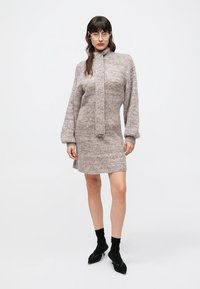 Πλεκτό φόρεμα σε ανοιχτό καφέ χρώμα με υφή, ψηλό γιακά και διακοσμητικό δέσιμο. Μακριά μανίκια και εφαρμοστή σιλουέτα. Φορεμένο με μαύρες κάλτσες και παπούτσια.