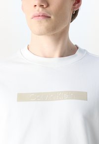 Felpa bianca in cotone con collo rotondo, collo elastico e un logo beige di Calvin Klein in rilievo sulla parte anteriore.