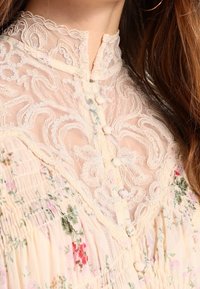 Světle květinová blůza s krajkovým průsvitem a vířivými vzory, zapínáním na knoflíky, jemnými texturami a měkkým krémovým pozadím.