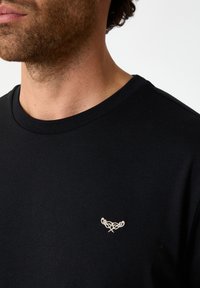 Black cotton t-shirt with a round neckline and a small white embroidered logo on the left chest. Smooth texture, casual design.