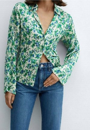Frau trägt eine grün-blaue, blumige, langärmelige Bluse mit Knopfverschluss, kombiniert mit hoch geschnittenen, blauen Jeans, die Hand auf der Taille.