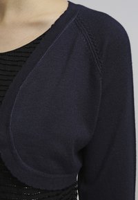 Marinblå stickad bolerokardigan med rundad framdesign, med texturerad söm längs ärmarnas och halsringningens kant.