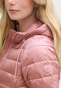 Heledat roosa puffer-jakk kapuutsiga, millel on tikandid, paelaga sulgemine ja ümmargune logo patch vasakul õlal.