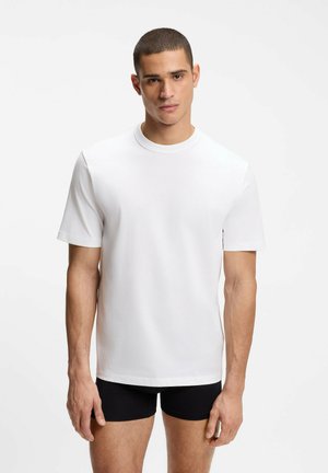 Junger Mann trägt ein schlichtes weißes Kurzarm-T-Shirt und schwarze Boxershorts und steht vor einem weißen Hintergrund.