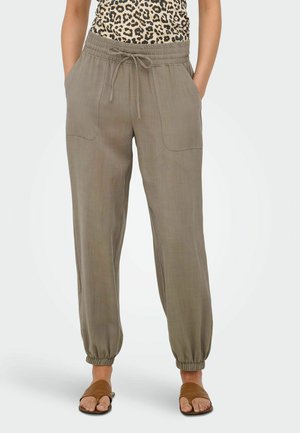 Vrouw die bruine linnen joggerbroek met trekkoord, een top met luipaardprint en bruine open-toe sandalen draagt, staand met handen in de zakken.