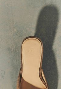 Svetlohnedá semišová obuv typu slip-on s polstrovanou stielkou vyrazeným nápisom „THE SET“ na textúrovanej šedej ploche vrhajúcej tieň.