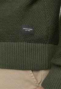 Pull en maille texturée vert foncé avec ourlet côtelé, présentant une étiquette noire portant la mention "JACK & JONES EST. 1990" en texte blanc.