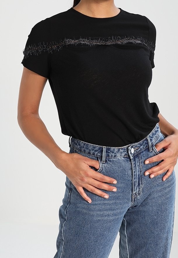 Mujer vestida con una blusa negra de manga corta con detalles deshilachados en el pecho, metida en unos vaqueros azul de cintura alta, con las manos apoyadas en las caderas.