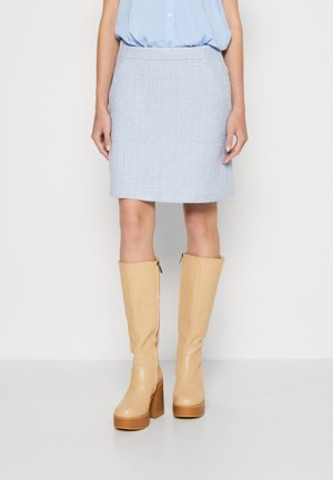 Vrouw draagt een lichtblauwe rok, bijpassende blouse en beige plateau-laarzen tot net onder de knie, staand tegen een effen achtergrond.