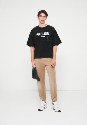 Czarna, oversized bawełniana koszulka z białym napisem, jasnobrązowe spodenki z weluru, białe sneakersy i trzymająca czarną czapkę. Minimalistyczne tło.
