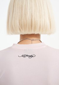 Světle růžové tričko s vyšitým černým logem "Ed Hardy" na zadní straně. Hladká bavlněná tkanina s kulatým výstřihem a standardním střihem.