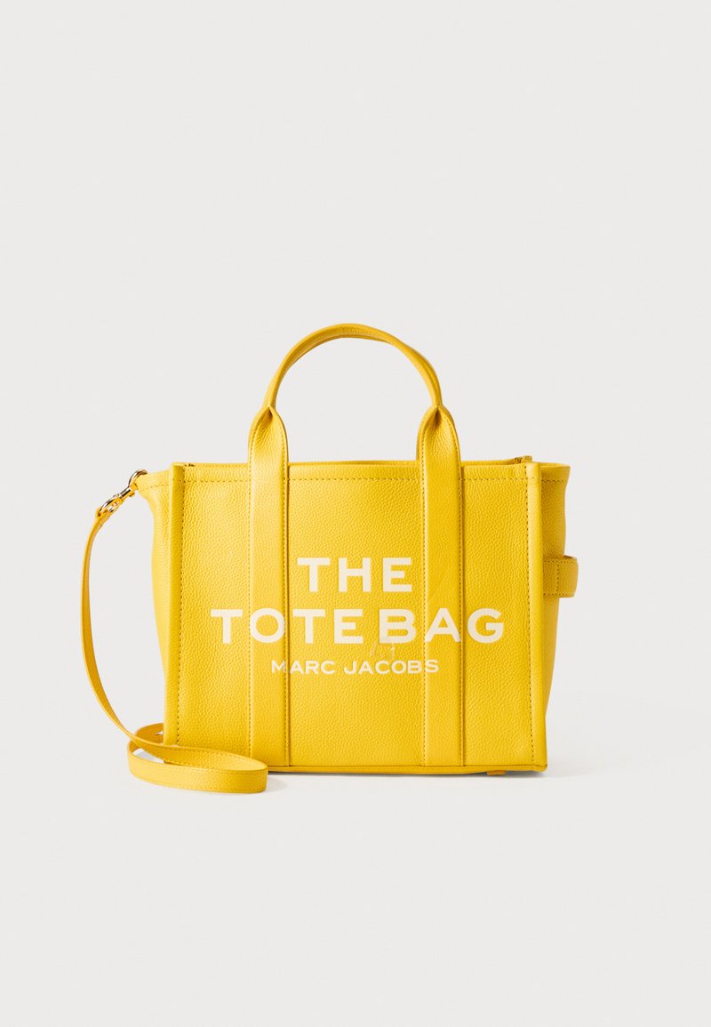 Yellow pebbled leather tote bag with dual handles, detachable shoulder strap, and bold white text: "THE TOTE BAG MARC JACOBS" on the front.