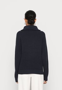 Pull overdimensionné marine en maille côtelée, avec un col haut, des épaules tombantes et des manches larges. Associé à un pantalon de couleur claire.