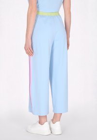 Világoskék, bő szárú nadrág textúrált zöld elasztikus derékkal és rózsaszín oldalsávokkal, sima anyagból készült. Fehér tornacipők láthatóak.