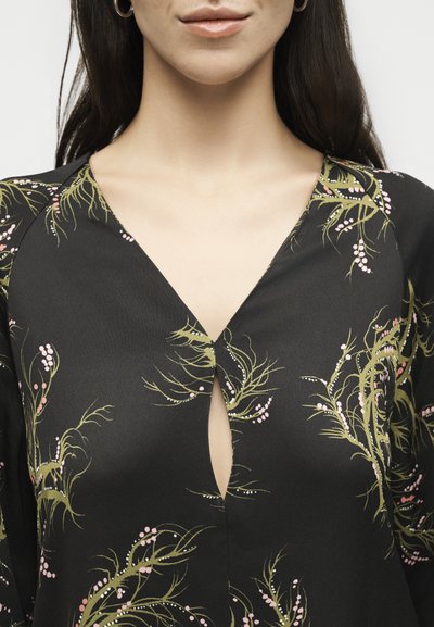 Γυναίκα που φοράει μαύρο μπλουζάκι με V λαιμόκοψη και πράσινο και ροζ βοτανικό σχέδιο, εμφανίζοντας μερικώς τον λαιμό, το πηγούνι και τα χείλη.