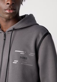 Felpa grigia con zip e dettagli neri, caratterizzata da un logo testurizzato e un motivo lineare sul petto. Materiale morbido, spessore medio.