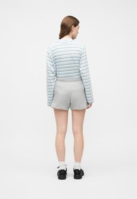 Γυναίκα που στέκεται με την πλάτη προς την κάμερα, φορώντας μπλε και άσπρη ριγέ μακρυμάνικη μπλούζα, γκρι σορτς, λευκές κάλτσες και μαύρα παπούτσια.