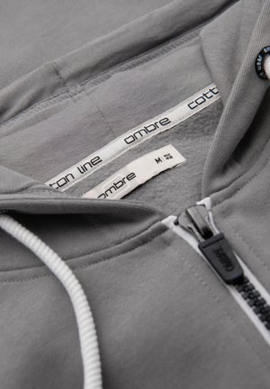 Sudadera con capucha gris con cremallera, cordones blancos visibles, cremallera etiquetada con "ombre" y etiquetas interiores que muestran la talla M EUR 48 y detalles de la marca.