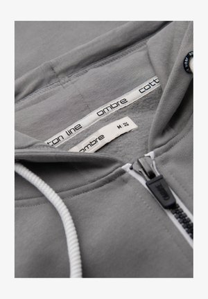 Sudadera con capucha gris con cremallera, cordones blancos visibles, cremallera etiquetada con "ombre" y etiquetas interiores que muestran la talla M EUR 48 y detalles de la marca.
