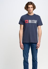 Jūras zils kokvilnas T-krekls ar sarkanu kvadrātu un baltu tekstu "BIG STAR", apaļa kakla izgriezums, īsas piedurknes, valkāts ar zilām džinsām.