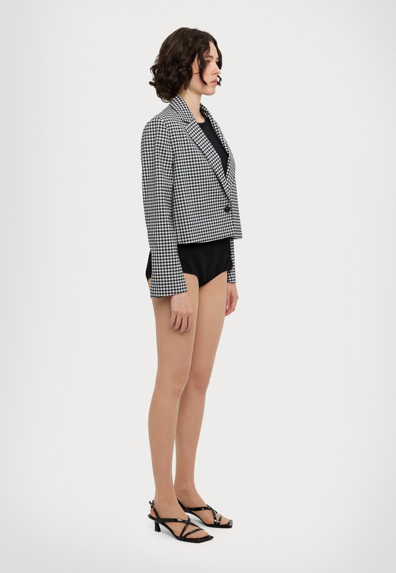 Blazer cropped a scacchi in bianco e nero con un bottone, indossato sopra una maglietta nera. Abbinato a sandali con tacco a strappo neri, vista laterale.