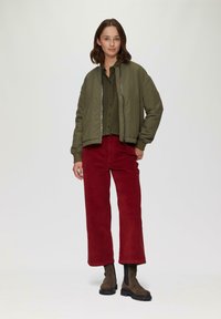 Veste bomber vert olive, chemise kaki boutonnée, pantalon en velours côtelé rouge à large jambe, et bottines marron avec semelles noires.