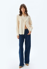 Crème-kleurige button-up shirt met rafelige details, gecombineerd met wijde donkerblauwe denim jeans met franjes aan de onderkant.