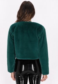 Afskåret teal faux fur-jakke med lange ærmer og blød tekstur. Kombineret med skinnende sorte bukser, hvilket skaber et kontrastfyldt look.