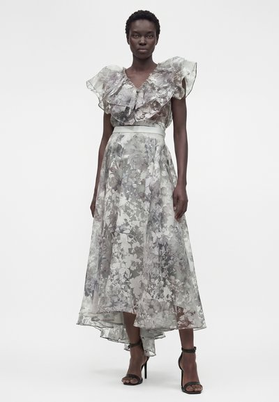 Γυναίκα φορεί ένα αμάνικο, γκρι και λευκό φόρεμα με λουλουδάτο μοτίβο, με βολάν V-λαιμόκοψη και ασύμμετρο τελείωμα, συνδυασμένο με μαύρα σανδάλια με ψηλό τακούνι.