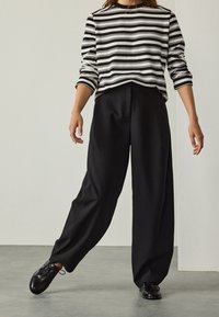 Persona che indossa pantaloni neri a gamba larga, scarpe nere con lacci e una maglia a maniche lunghe a righe bianche e nere in piedi su un pavimento grigio.