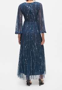 Mörkblå golvlång klänning med genomskinliga ärmar, dekorerad med paljetter i snirliga mönster, och en figurnära bodice som betonar midjan.