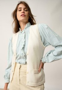 Biała wełniana kamizelka z pięcioma guzikami, noszona na jasnoniebieskiej koszuli z falbanami. Strój zawiera strukturę, paskowaną spódnicę. Neutralne tło.