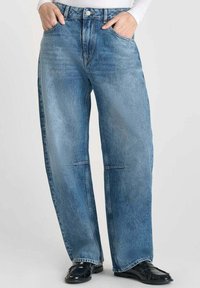 Personne portant un jean en denim bleu ample et des mocassins noirs, les mains dans les poches, se tenant contre un fond clair.