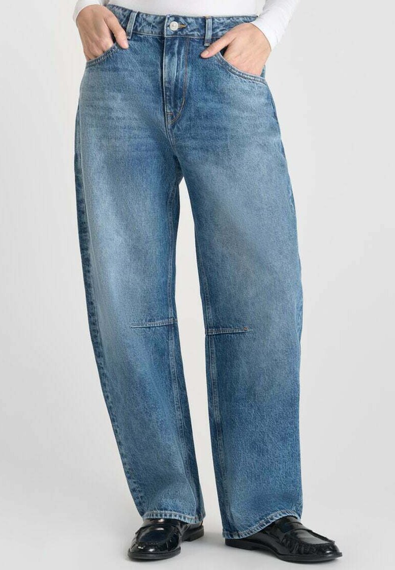 Personne portant un jean en denim bleu ample et des mocassins noirs, les mains dans les poches, se tenant contre un fond clair.