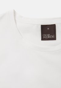 T-shirt bianco in cotone con scollatura rotonda. Presenta un'etichetta in tessuto marrone con "OSCAR JACOBSON" e taglia "M" ricamata sul retro.
