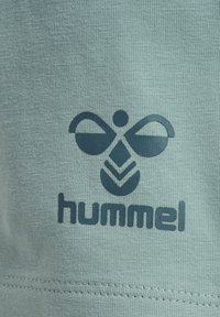 Ljust grönt tyg med en texturerad yta har en mörkblå "hummel"-logotyp och en ikon som liknar vingar samt ett bihuvud.