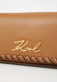 Hnědá syntetická kožená peněženka s vyraženým zlatým logem "Karl" a dekorativním prošíváním podél okraje. Hladký povrch a elegantní design.