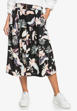 Femme portant une jupe fleurie noire mi-longue avec des boutons, des baskets blanches et un haut blanc à manches longues avec les manches retroussées.