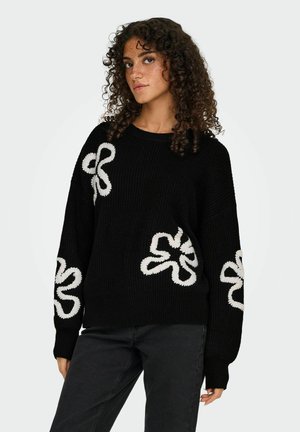 Zwarte gebreide trui met een oversized pasvorm, witte bloemenborduursels aan beide zijkanten en geribde manchetten. De textuur van de stof voegt diepte toe.
