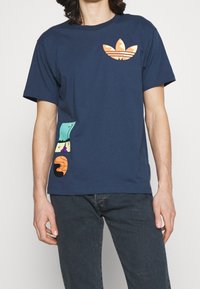 Námořnická modrá bavlněná tričko s oranžovým logem na hrudi a barevnými grafickými akcenty na straně. Krátké rukávy, volný střih.