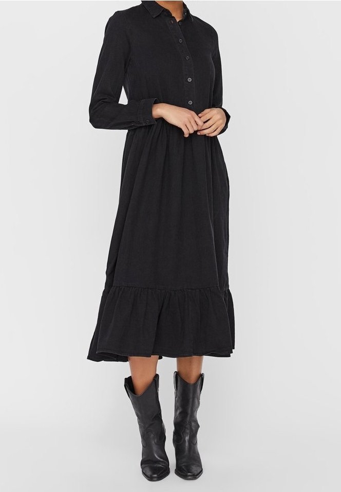 Femme portant une robe midi noire à manches longues boutonnée avec un ourlet à volants et des bottes en cuir noires mi-mollet, debout sur un fond gris.
