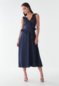 Laivastonsininen midi-mekko, jossa on röyhelöyksityiskohtia kaula-aukolla ja epäsymmetrinen helma, jossa on teksturoitu kangas ja istuva rumpu.