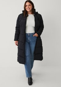 Zwarte pufferjas met een ritssluiting aan de voorkant, geplooide mouwen en een capuchon. Heeft gewatteerde secties en een middellange snit, gecombineerd met blauwe jeans en laarzen.