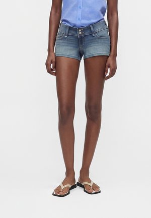 Persoon draagt lichtblauw overhemd met knopen, denim shorts met dubbele knopen en beige sandalen met zwarte zolen, staande op witte achtergrond.