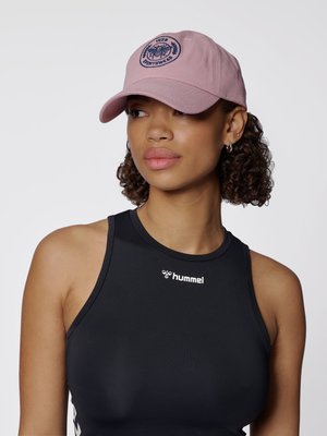 Vrouw met krullend haar die een roze pet met een logo draagt en een zwarte mouwloze sporttop, licht naar opzij kijkend.