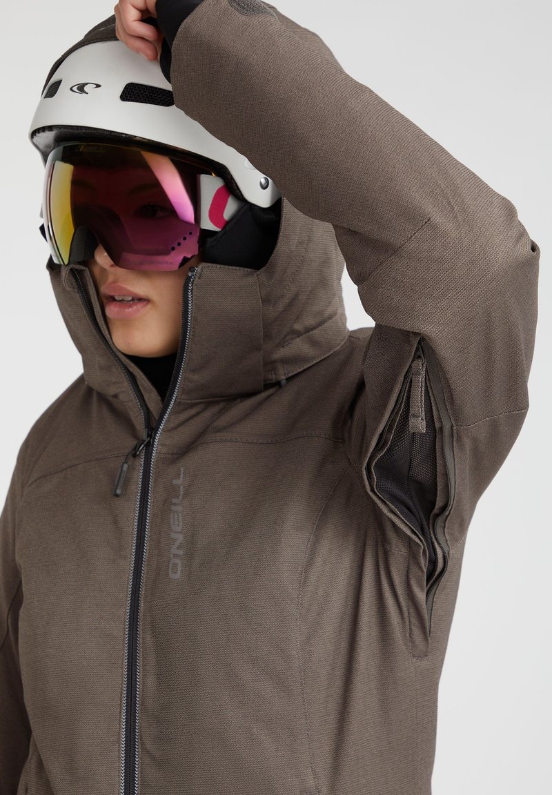 Braune isolierte Jacke mit hohem Kragen, Reißverschluss-Taschen und subtiler Textur. Getragen mit einem weißen Helm und getönten Goggles.