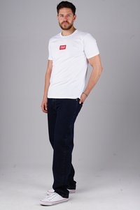 Weißes T-Shirt mit rotem rechteckigem Logo, kombiniert mit dunkelblauen Jeans und weißen Sneakern mit roten und blauen Akzenten.